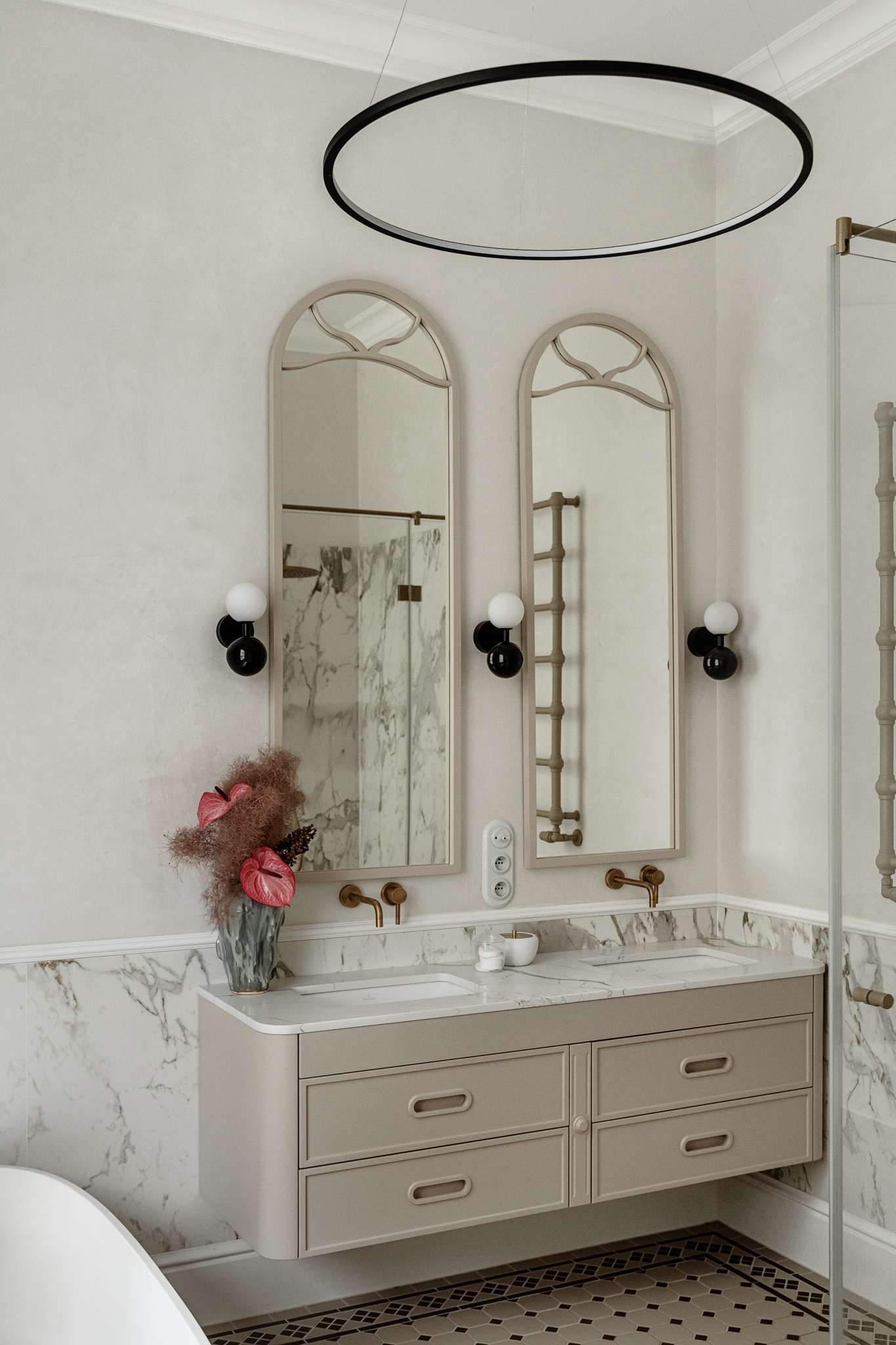 In the bathroom, classic materials and soft curves create a serene, gallery-like atmosphere. Arched mirrors nod to the apartment’s Art Nouveau heritage, while brass fixtures, decorative Ottocento plaster walls,  and a floating taupe vanity bring in modern refinement. A sculptural tub with a matte finish sits beneath a sunlit window, framed by Calacatta Gold marble walls and warm metal accents.