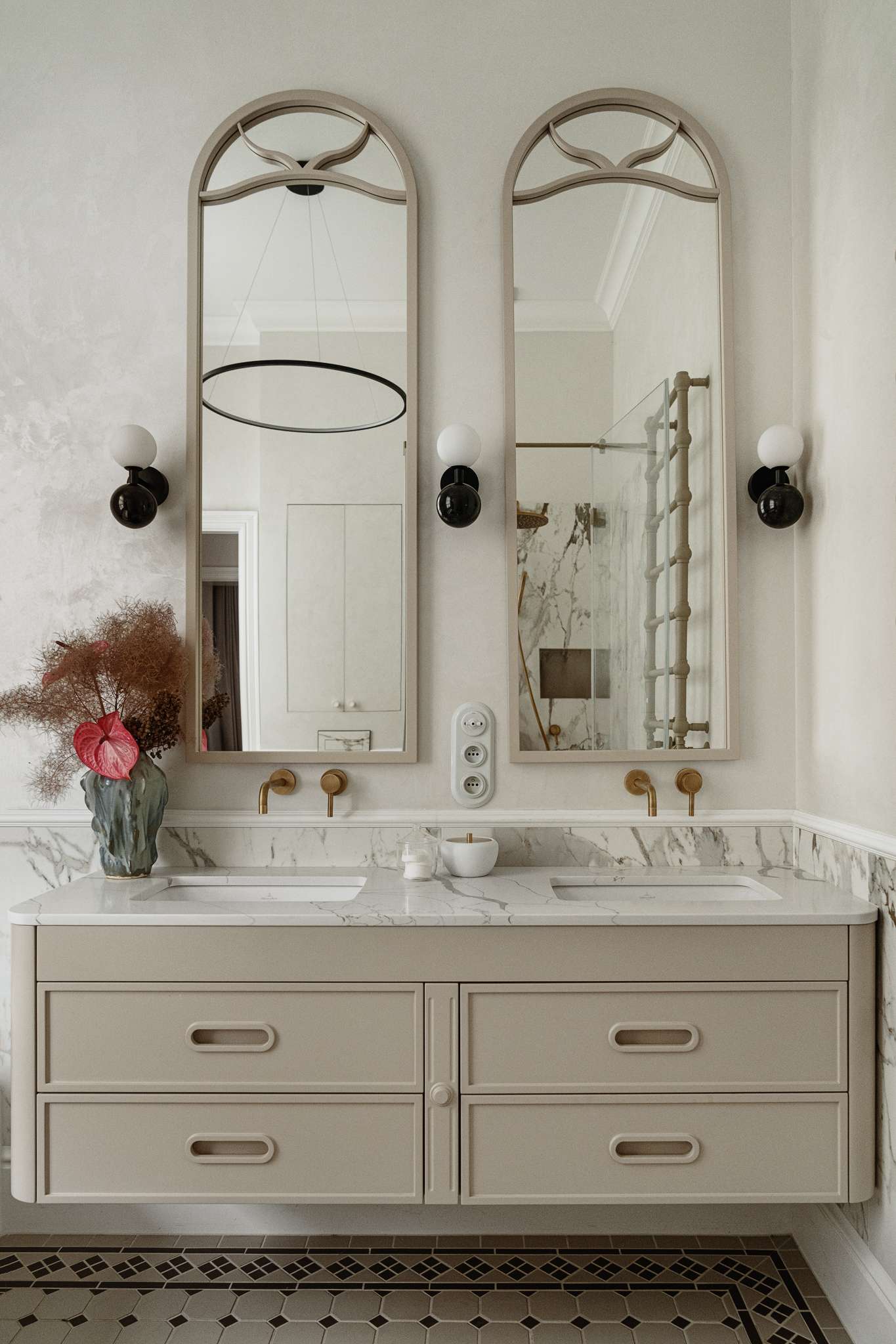 In the bathroom, classic materials and soft curves create a serene, gallery-like atmosphere. Arched mirrors nod to the apartment’s Art Nouveau heritage, while brass fixtures, decorative Ottocento plaster walls,  and a floating taupe vanity bring in modern refinement. A sculptural tub with a matte finish sits beneath a sunlit window, framed by Calacatta Gold marble walls and warm metal accents.