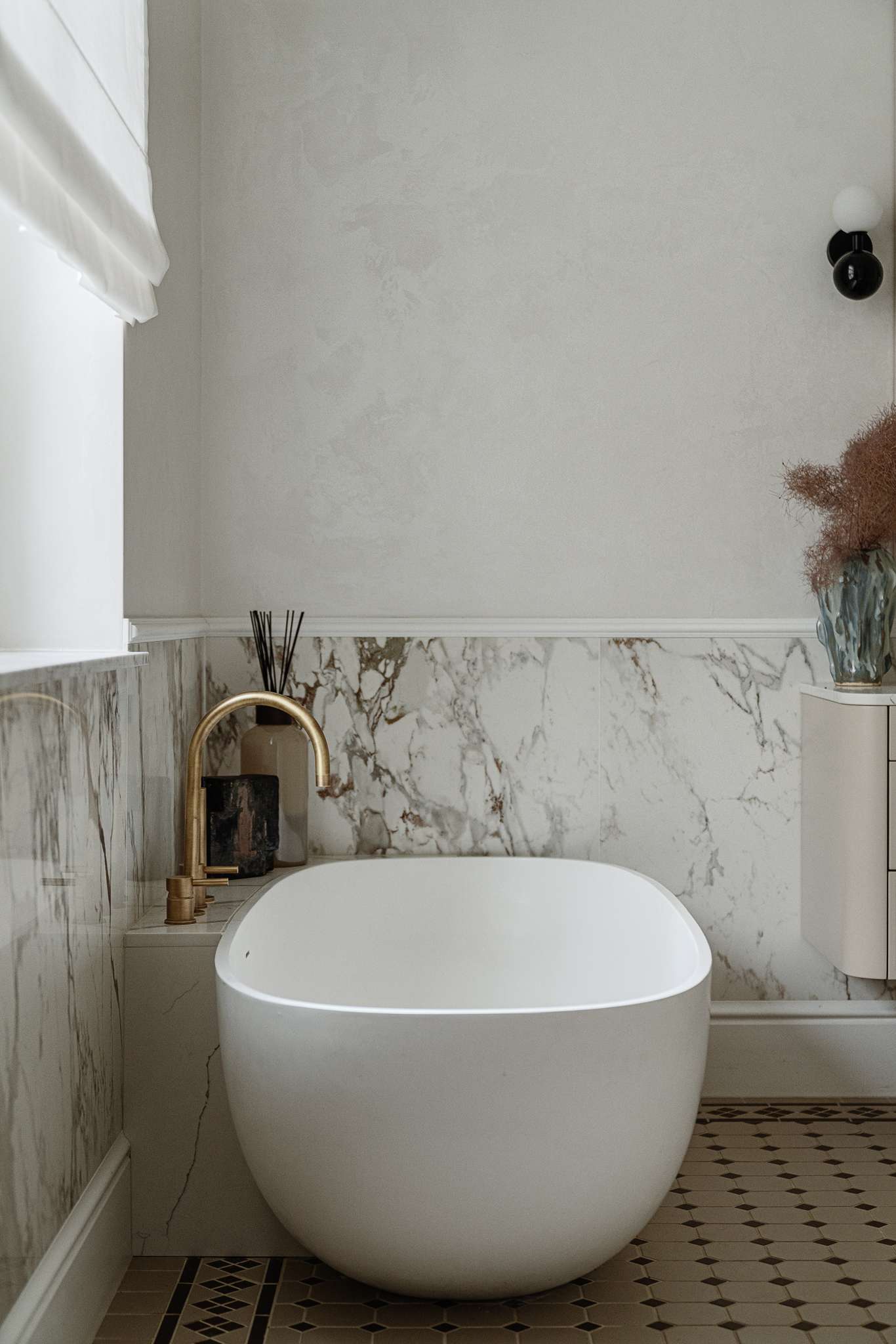 In the bathroom, classic materials and soft curves create a serene, gallery-like atmosphere. Arched mirrors nod to the apartment’s Art Nouveau heritage, while brass fixtures, decorative Ottocento plaster walls,  and a floating taupe vanity bring in modern refinement. A sculptural tub with a matte finish sits beneath a sunlit window, framed by Calacatta Gold marble walls and warm metal accents.
