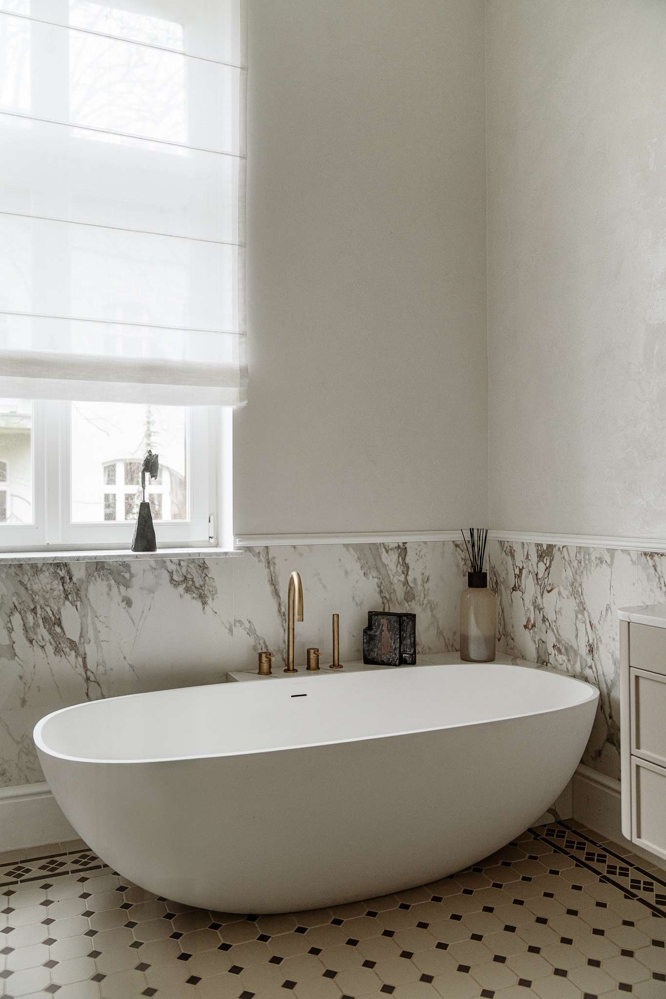 In the bathroom, classic materials and soft curves create a serene, gallery-like atmosphere. Arched mirrors nod to the apartment’s Art Nouveau heritage, while brass fixtures, decorative Ottocento plaster walls,  and a floating taupe vanity bring in modern refinement. A sculptural tub with a matte finish sits beneath a sunlit window, framed by Calacatta Gold marble walls and warm metal accents.