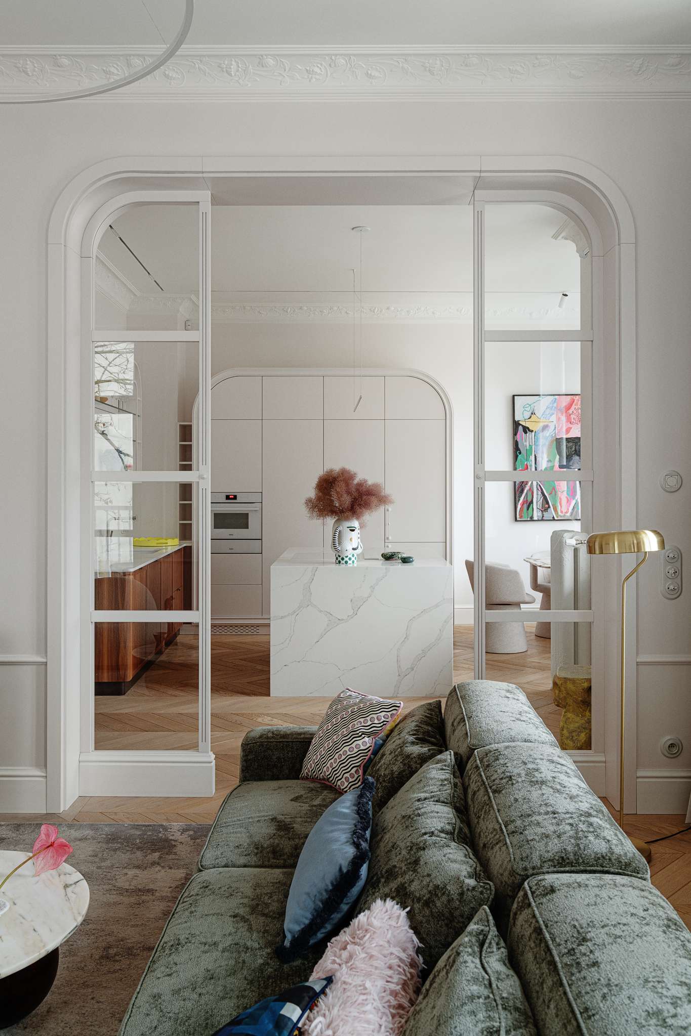 This kitchen pairs sculptural softness with practical sophistication. A central island in marble-look stone anchors the space, its clean lines echoed in the curved cabinetry and arched built-ins. Walnut lower cabinets add warmth and grain, while open shelving displays a curated mix of ceramics and glassware. 