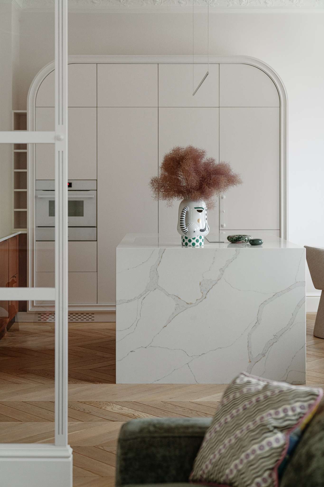 This kitchen pairs sculptural softness with practical sophistication. A central island in marble-look stone anchors the space, its clean lines echoed in the curved cabinetry and arched built-ins. Walnut lower cabinets add warmth and grain, while open shelving displays a curated mix of ceramics and glassware. 