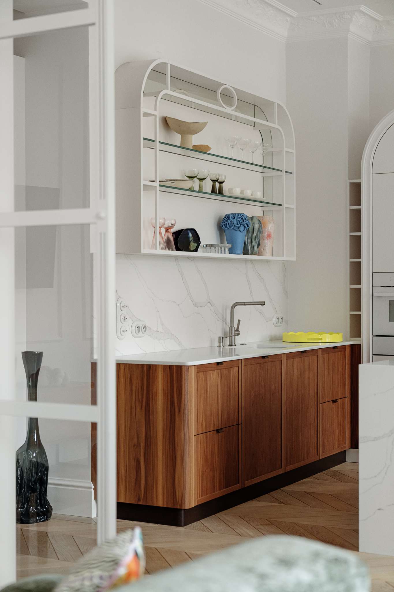 This kitchen pairs sculptural softness with practical sophistication. A central island in marble-look stone anchors the space, its clean lines echoed in the curved cabinetry and arched built-ins. Walnut lower cabinets add warmth and grain, while open shelving displays a curated mix of ceramics and glassware. 