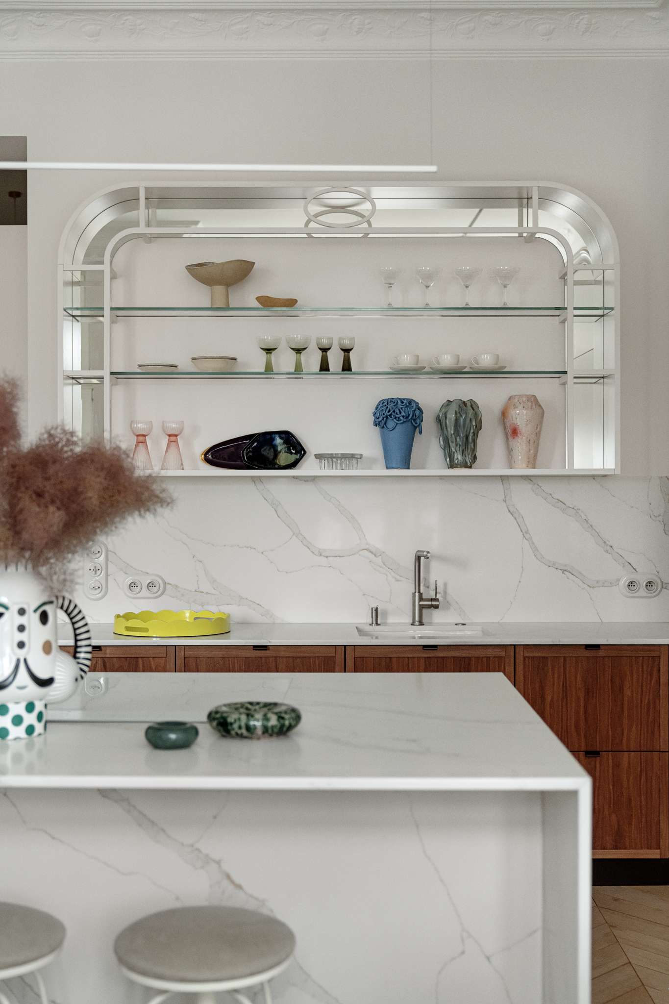 This kitchen pairs sculptural softness with practical sophistication. A central island in marble-look stone anchors the space, its clean lines echoed in the curved cabinetry and arched built-ins. Walnut lower cabinets add warmth and grain, while open shelving displays a curated mix of ceramics and glassware. 