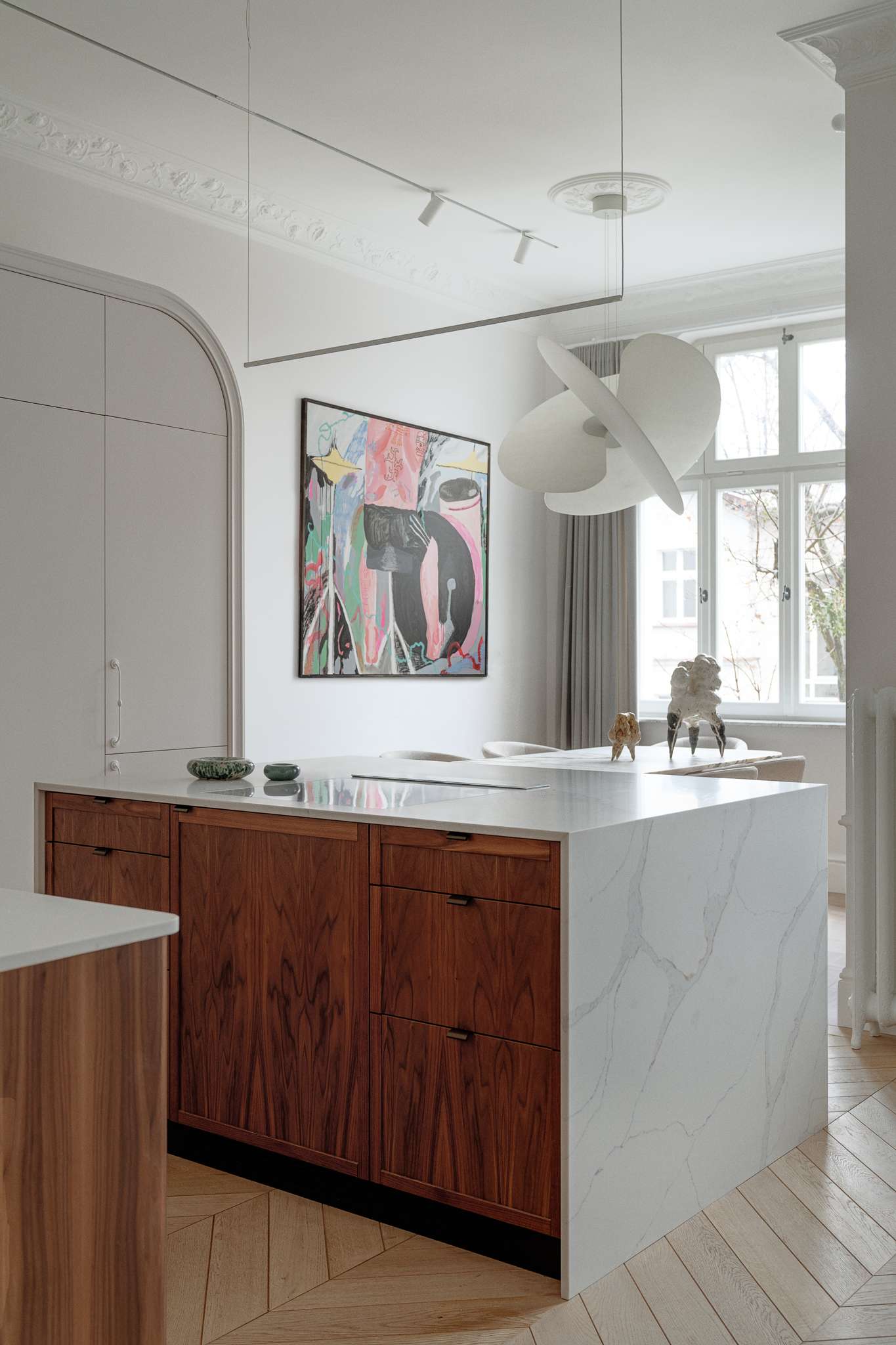 This kitchen pairs sculptural softness with practical sophistication. A central island in marble-look stone anchors the space, its clean lines echoed in the curved cabinetry and arched built-ins. Walnut lower cabinets add warmth and grain, while open shelving displays a curated mix of ceramics and glassware. 