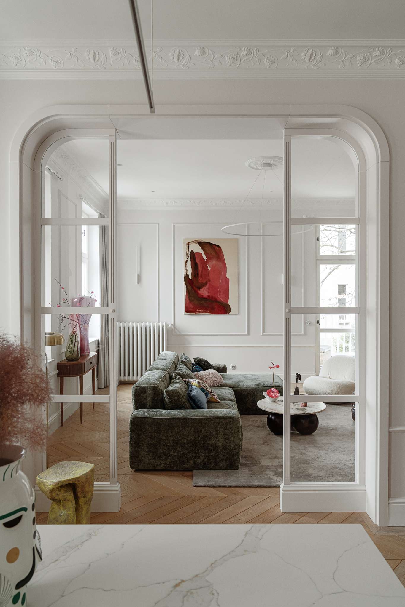 This living room combines refined elegance with cozy textures and bold accents. A plush green sectional anchors the space, softened by layered cushions in playful prints and hues. The marble-topped coffee table rests on rounded spheres, grounding the room with sculptural weight. A curved white lounge chair and a deep-toned side table add contrast and shape, while a vibrant pink abstract painting by Aleksandra Szewczuk brings warmth to the pale walls. 