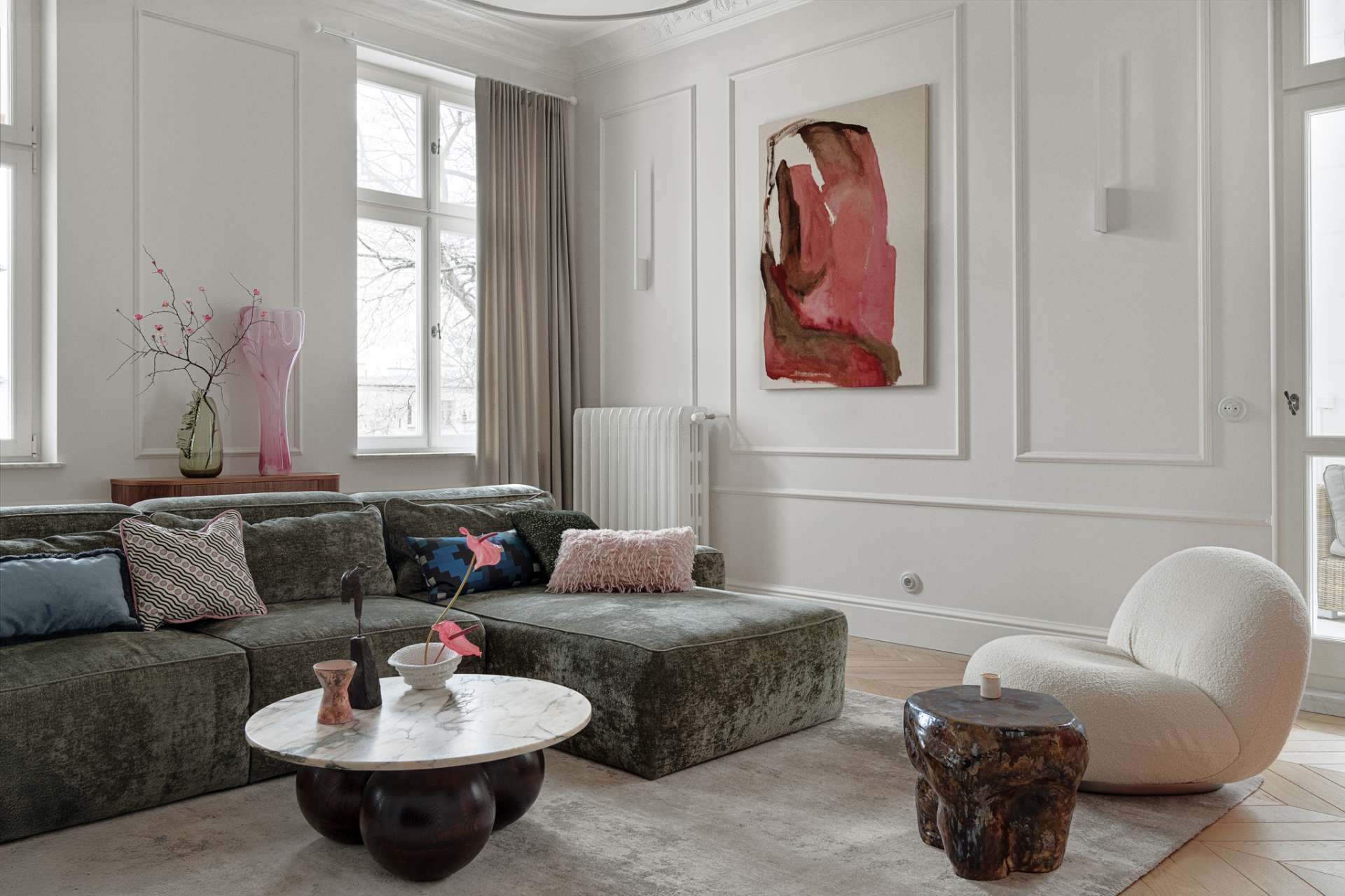 This living room combines refined elegance with cozy textures and bold accents. A plush green sectional anchors the space, softened by layered cushions in playful prints and hues. The marble-topped coffee table rests on rounded spheres, grounding the room with sculptural weight. A curved white lounge chair and a deep-toned side table add contrast and shape, while a vibrant pink abstract painting by Aleksandra Szewczuk brings warmth to the pale walls. 