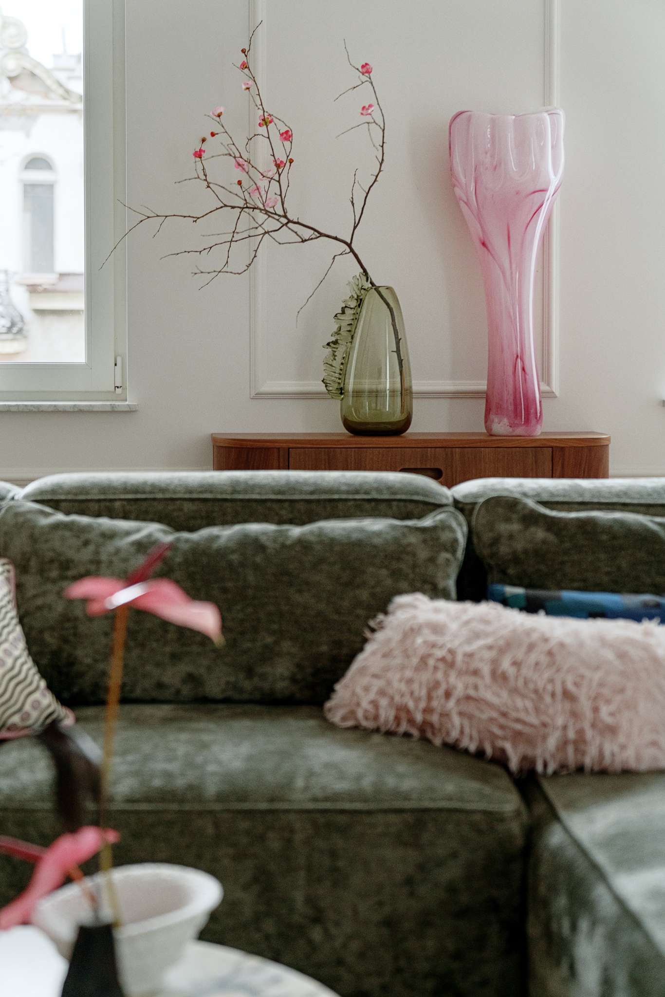 This living room combines refined elegance with cozy textures and bold accents. A plush green sectional anchors the space, softened by layered cushions in playful prints and hues. The marble-topped coffee table rests on rounded spheres, grounding the room with sculptural weight. A curved white lounge chair and a deep-toned side table add contrast and shape, while a vibrant pink abstract painting by Aleksandra Szewczuk brings warmth to the pale walls. 