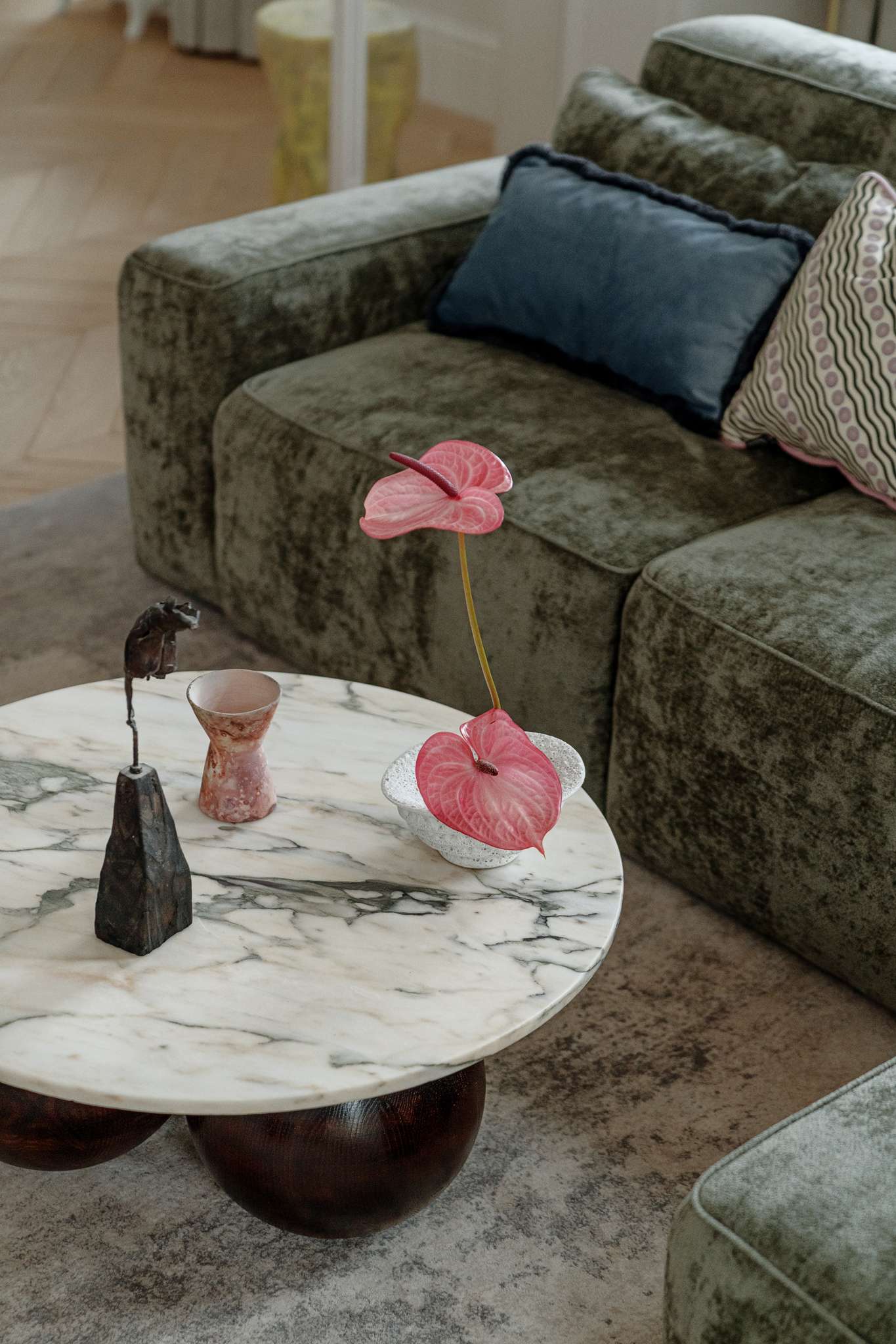 This living room combines refined elegance with cozy textures and bold accents. A plush green sectional anchors the space, softened by layered cushions in playful prints and hues. The marble-topped coffee table rests on rounded spheres, grounding the room with sculptural weight. A curved white lounge chair and a deep-toned side table add contrast and shape, while a vibrant pink abstract painting by Aleksandra Szewczuk brings warmth to the pale walls. 
