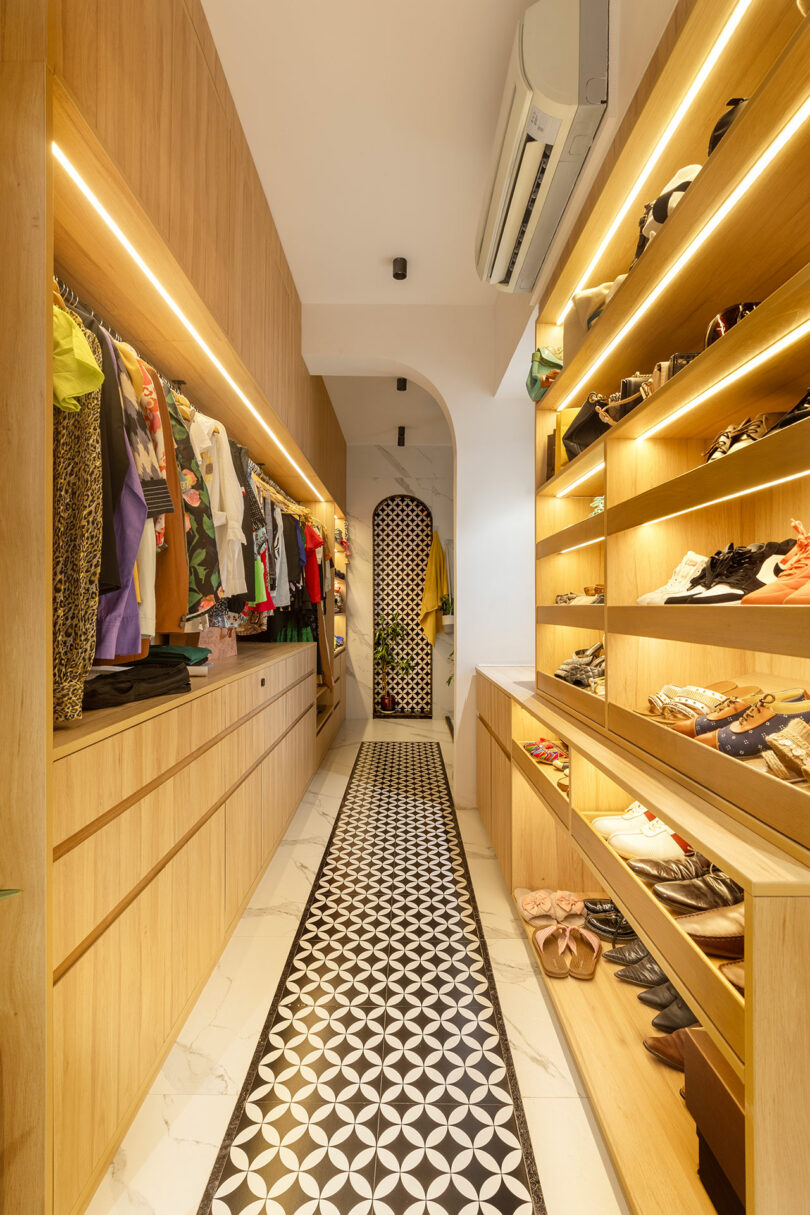 A narrow, well-lit walk-in closet features wooden shelves with shoes, drawers, hanging clothes, a patterned tile floor runner, and an air conditioner on the ceiling.