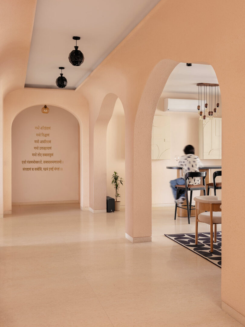 A hallway with peach-colored walls and arches, wall text in Hindi, a small plant, and a blurred person sitting at a table in a modern, minimalist interior.